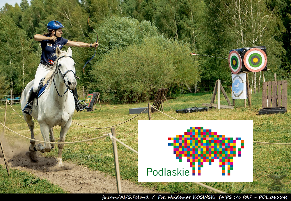 Po lewej stronie zawodniczka Anna Sokólska na białym koniu w galopie strzela z łuku do celu, w prawym dolnym rogu logo kolorowego żubra, partner Województwo Podlaskie